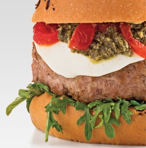 Bun topped with arugula, burger patty, melted cheese, pesto, and sliced red peppers