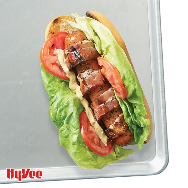 Grilled hot dog and bun topped with lettuce, tomatoes, bacon and mayo