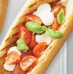 Grilled hot dog and bun topped with mozzarella pieces, sliced cherry tomatoes and basil leaves
