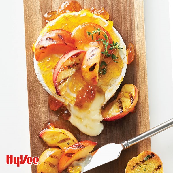 Wheel of brie topped with apricot preserves and grilled peaches and plums on a wooden board with a cheese knife