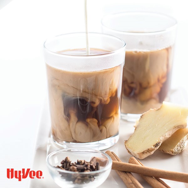 Creamer being pored into a clear glass of chi tea and a side of whole cloves, cinnamon sticks, and fresh ginger