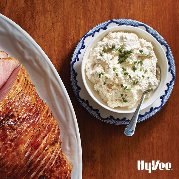 Whole sliced and glazed ham next to horseradish spread garnished with fresh chopped chives
