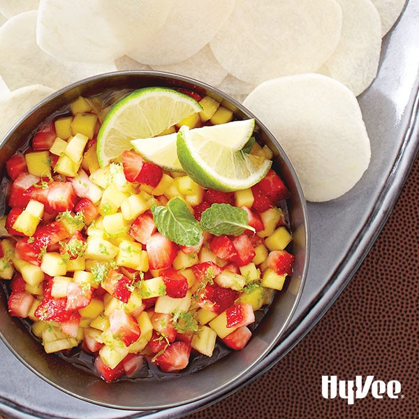 Metal bowl filled with chopped pineapple, strawberries, lime zest, lime wedges, and fresh mint leaves with a side of potato chips