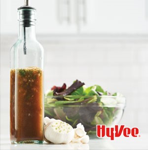 Glass olive oil bottle filled with herb vinaigrette next to whole clove garlic and mixed green salad