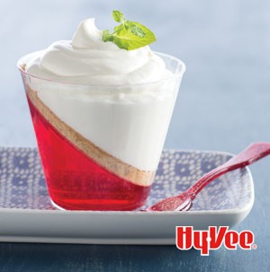 Glass filled with slanted layers of red gelatin, graham cracker crumbs, and creamy cheesecake filling