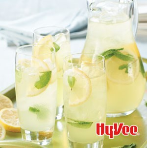 Three glasses of mint lemonade on a try with pitcher with sliced lemon wedges and mint leaves as garnish 