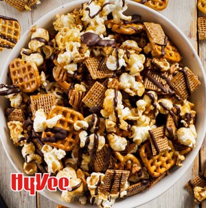 Bowl of mixed popcorn, cereal, pretzel squares and pecans, drizzled in dark chocolate