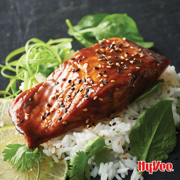 Glazed salmon topped with black sesame seeds over rice garnished with cilantro, lime slices, basil, and green onions