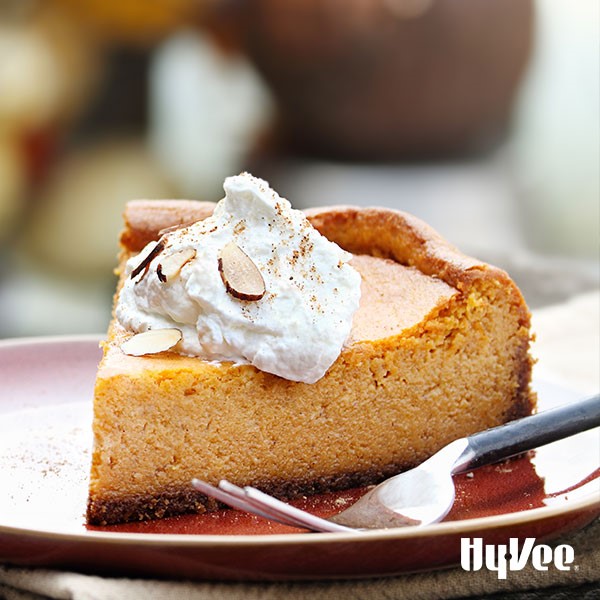 Slice of pumpkin pie on a red plate with a fork and a dollop of whipped topping with sliced almonds