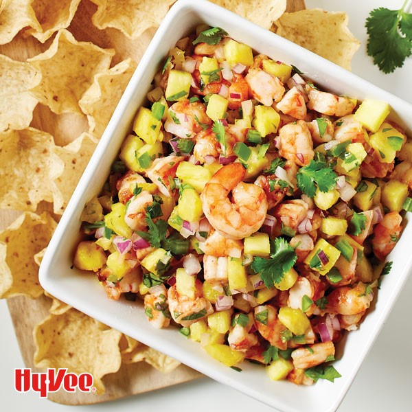 Cooked shrimp, diced pineapple, diced red onion, and pineapple in a square white dish surrounded by tortilla scoop chips