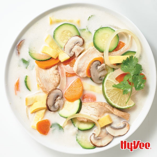 White bowl filled with white coconut soup with large pieces of sliced chicken, mushrooms, carrots, zucchini and garnished with lime wedge and fresh cilantro