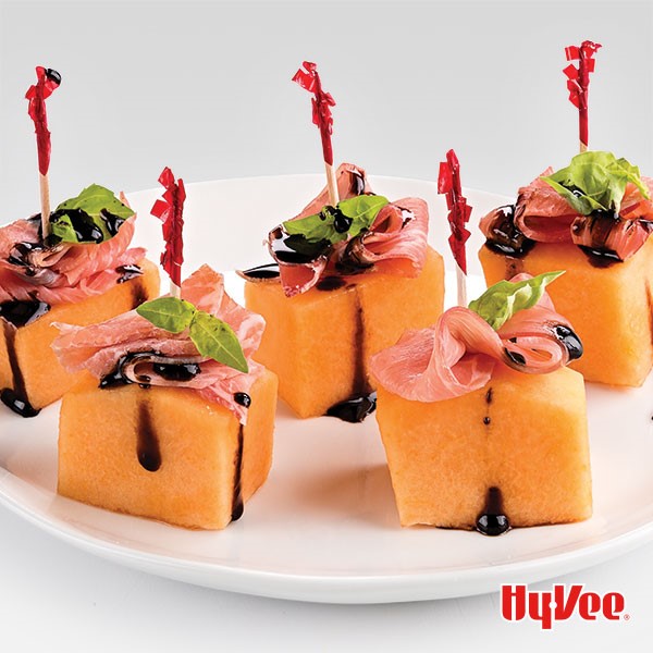 White plate of cantaloupe cubes topped with prosciutto, basil and balsamic and held together with a toothpick