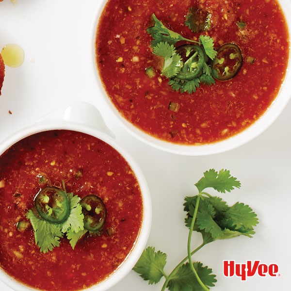 Two bowls full or red gazpacho and topped with sliced jalapenos and fresh cilantro