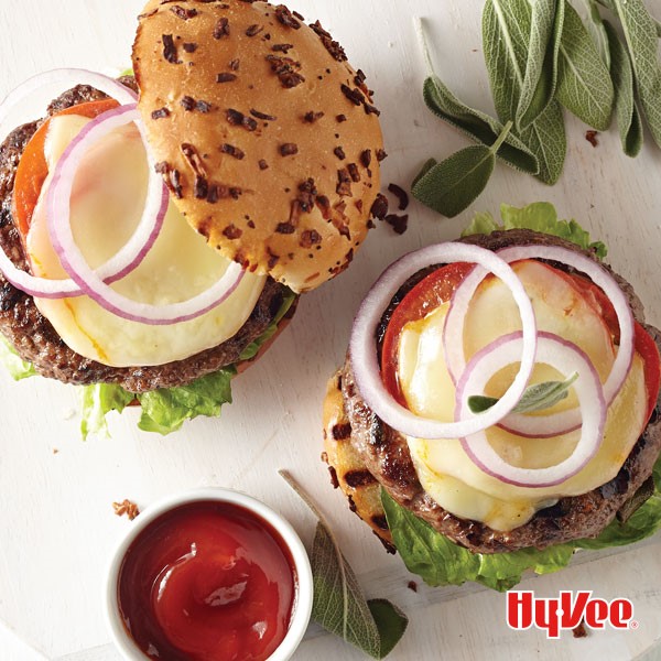 Grilled burgers on top of buns with sliced tomatoes, cheese, red onion rings, with sage leaves and ketchup on the side