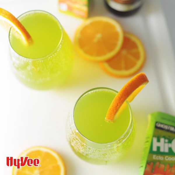 Lime green drinks in clear glasses topped with orange wedges with orange slices on the side