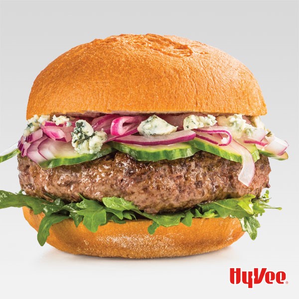 Bun topped with arugula, burger patty, sliced cucumbers, red onions, and crumbled Gorgonzola cheese 
