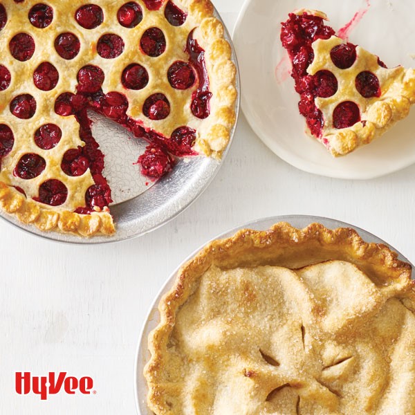 Cherry pie with circles cut out of top crust and a wedge on a plate next to double crusted pie with fluted crust