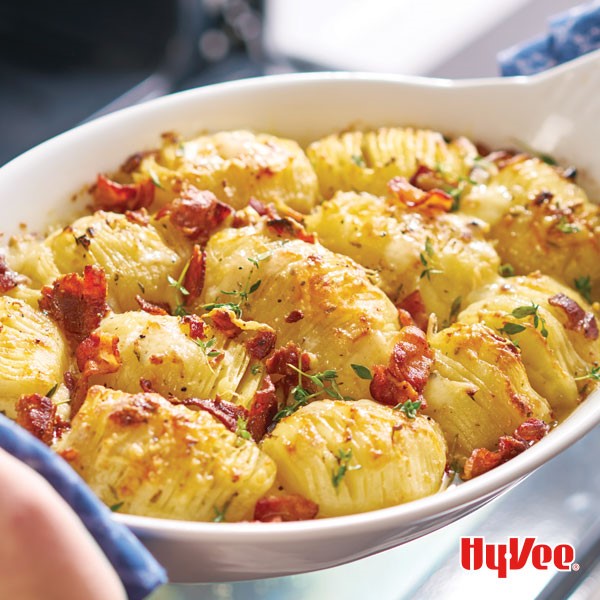 Hasselback potatoes in white casserole dish garnished with fresh thyme and cooked, chopped bacon