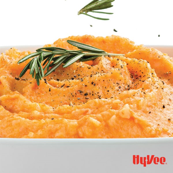 Orange vegetable mash in a casserole dish topped with black pepper and fresh rosemary sprigs