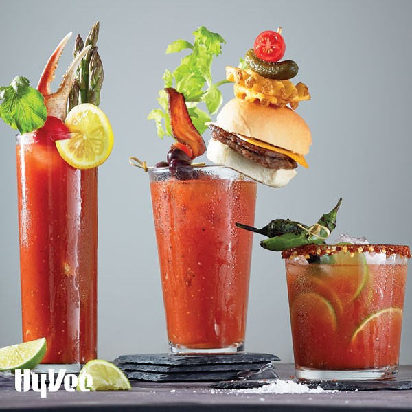 Three glasses filled with bloody marys and topped with crab claws, mini burgers, hot peppers, olives, and bacon