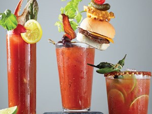 Three glasses filled with bloody marys and topped with crab claws, mini burgers, hot peppers, olives, and bacon