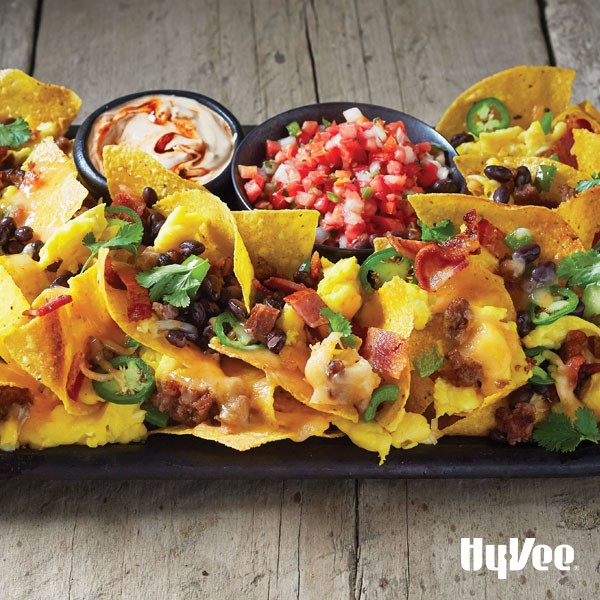 Plate of tortilla chips topped with bacon, black beans, sausage, green bell pepper, shredded cheese, green onion and cilantro, served with side of salsa and sour cream