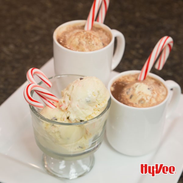 Peppermint ice cream in a glass with candy canes next to two mugs of hot chocolate