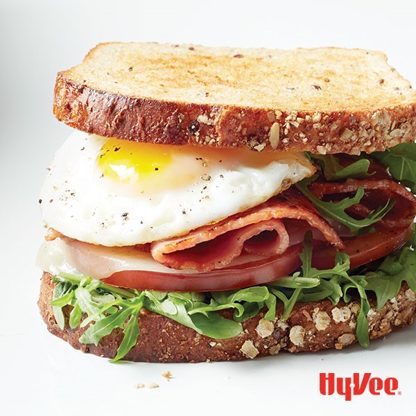 Sandwich filled with arugula, tomato slices, melted cheese, bacon, and a fried egg