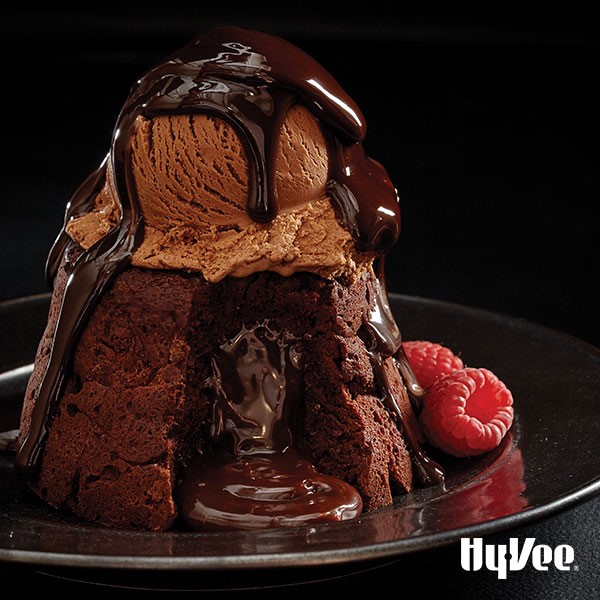 Molten lava cake split open with oozing chocolate topped with chocolate ice cream and hot fudge and garnished with fresh raspberries