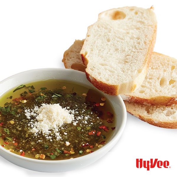 Stacked bread slices next to white bowl filled with olive oil, balsamic, red pepper flakes, grated Parmesan cheese, and sliced basil