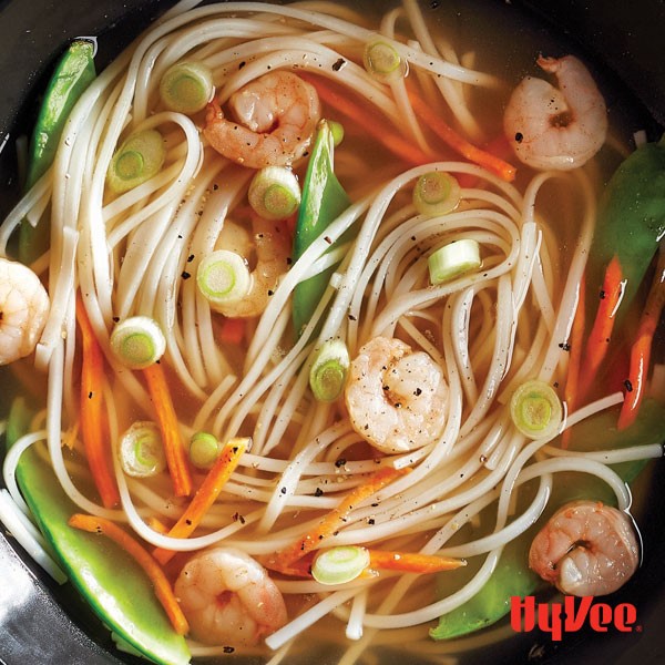 Pot of soup broth mixed with thin rice noodles, shredded carrots, shrimp, snow peas and green onions