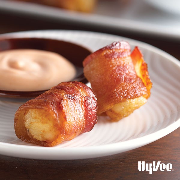 Tater tots wrapped in cooked bacon on white plate with dipping sauce in background