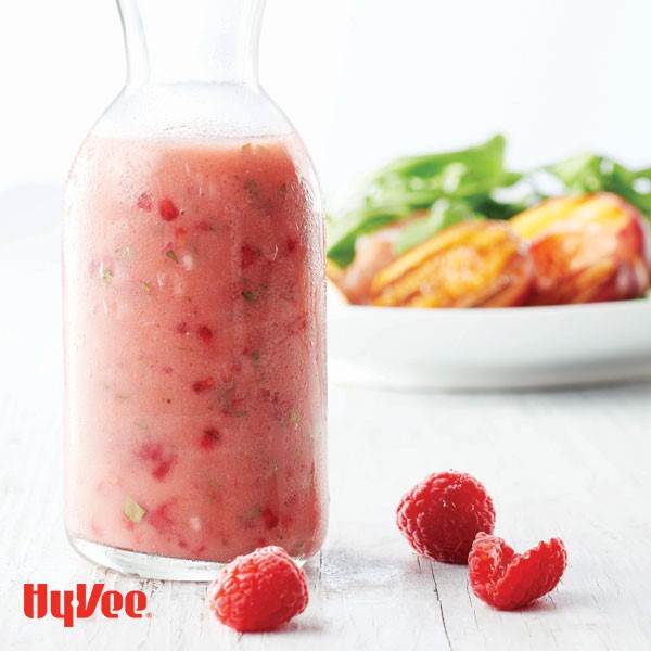 Raspberry vinaigrette in a tall glass container with fresh raspberries on the side and salad in background
