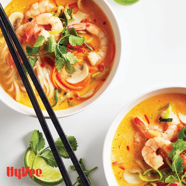 Orange broth filled with rice noodles, shredded carrots, sliced mushrooms, and tail-on cooked shrimp garnished with fresh cilantro