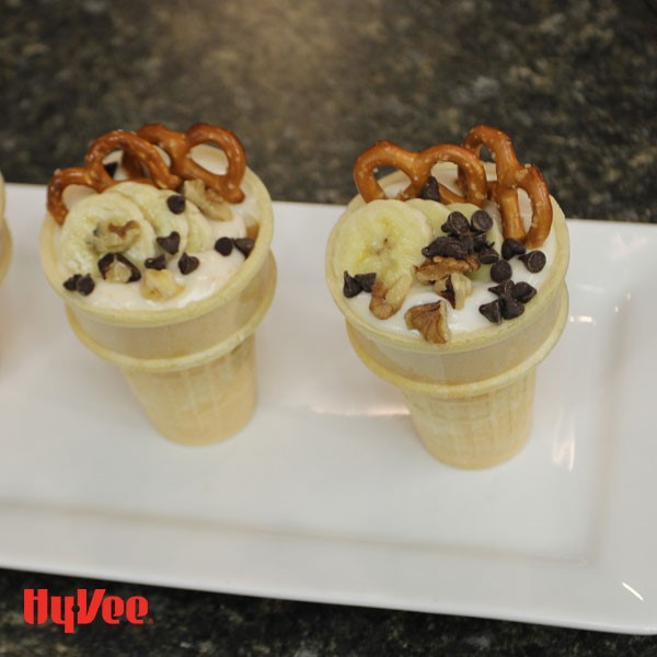 Plate of ice cream cones filled with yogurt, banana slices, walnuts, pretzles and mini chocolate chips 