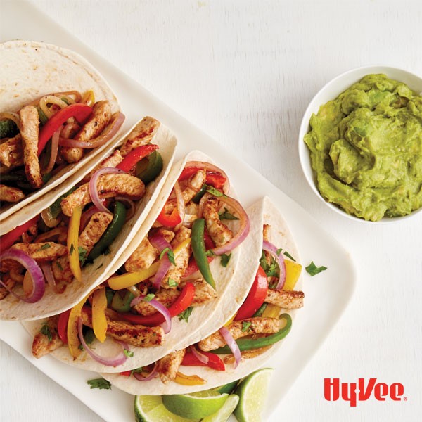 Fajitas in flour tortillas with sliced pork, bell peppers, onions, lime wedges, and side of guacamole