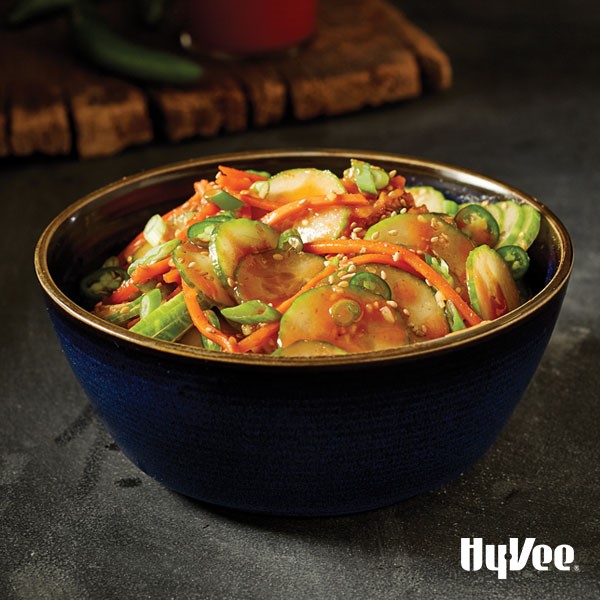 Kimchi in a blue bowl with sliced cucumbers, peppers and green onions with shredded carrots