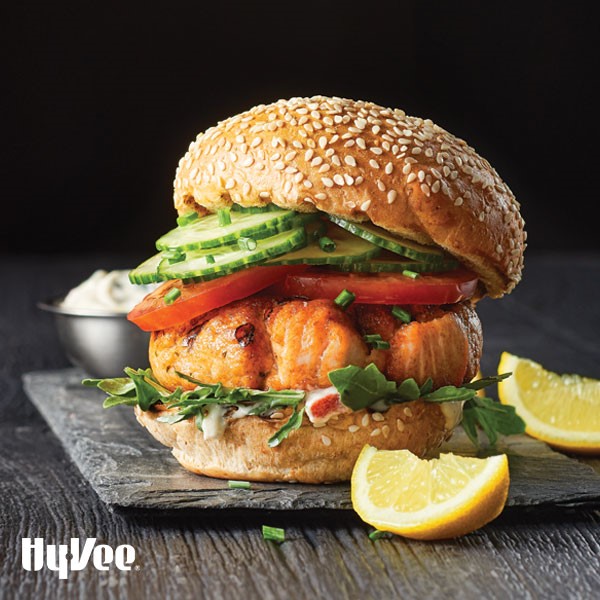 Bun topped with creamy sauce, arugula, salmon burger, sliced tomatoes, and sliced cucumbers with fresh lemon wedges as garnish