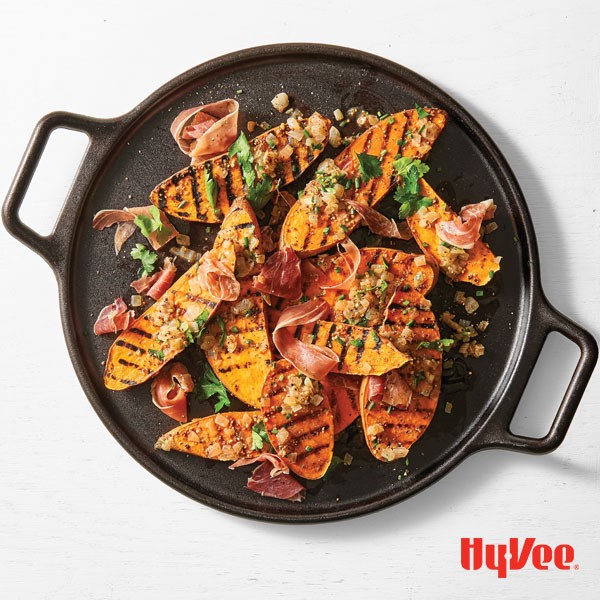 Black cast-iron pan filled with grilled sweet potato halves, prosciutto, and fresh herbs for garnish