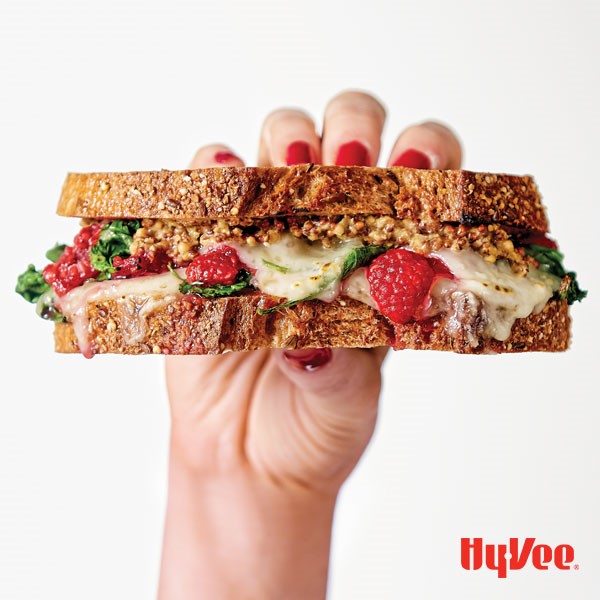Hand holding sandwich with melted cheese, spinach, whole grain mustard, and raspberries