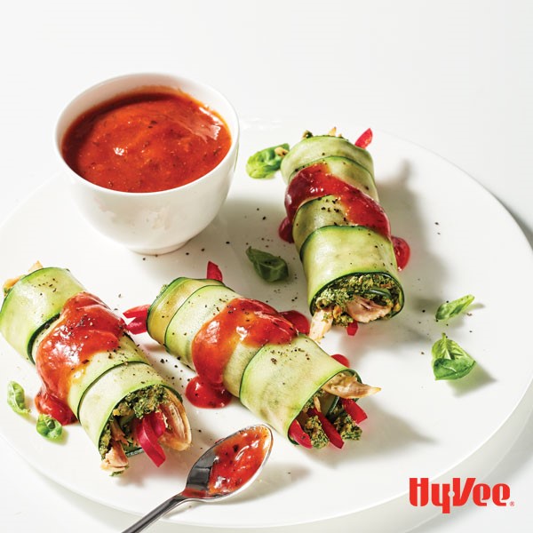 Thinly sliced zucchini wraps around various vegetables and is garnished with basil leaves and drizzled in red sauce