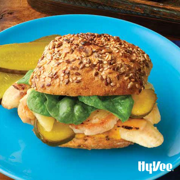 Blue plate with bun topped with pickles, spinach, grilled bananas, and meat on a seeded bun