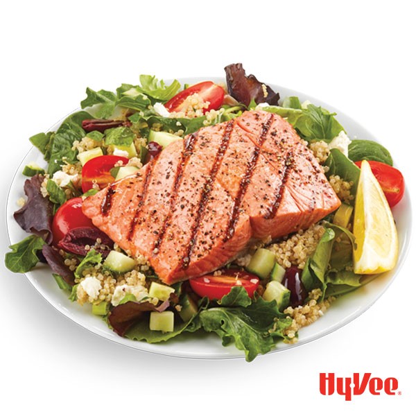 Grilled salmon atop mixed greens, diced cucumbers, halved cherry tomatoes, cooked quinoa, and garnished with fresh lemon wedge