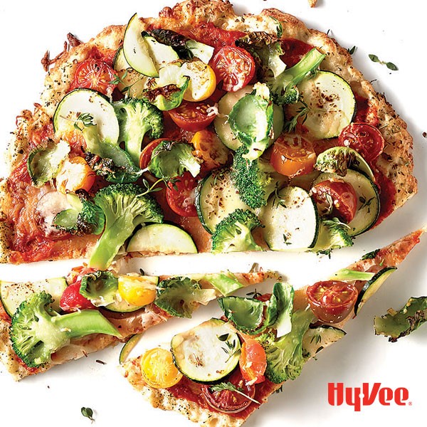 Wedged gluten-free pizza topped with halved red and yellow cherry tomatoes, sliced zucchini, brussel sprouts, and broccoli florets 