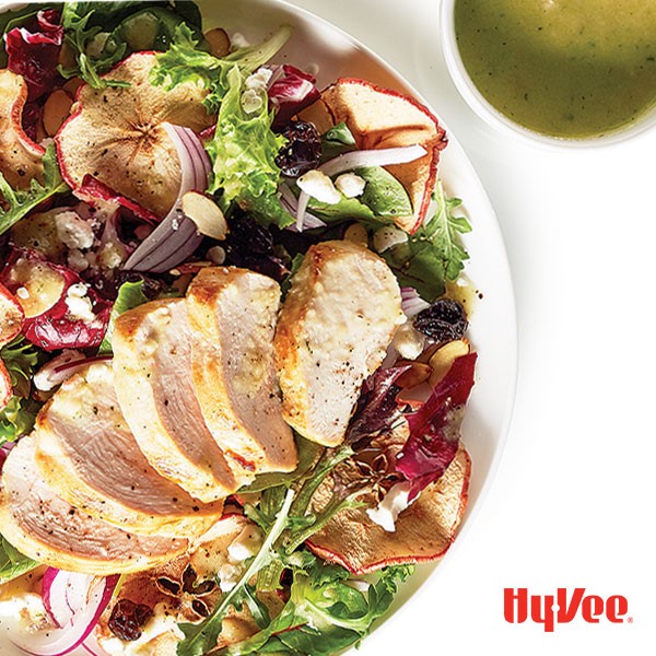 White bowl filled with oven-dried apple chips, sliced red onions, crumbled cheese, mixed greens and sliced cooked chicken