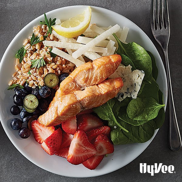 White plate filled with spinach, sliced strawberries, blueberries, farro, sliced jicama, two pieces of salmon, and sliced Gorgonzola cheese