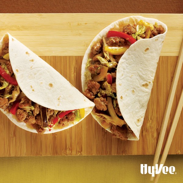 Flour tortillas wrapped around sauteed vegetables and ground cooked pork on a wooden cutting board with wooden chopsticks