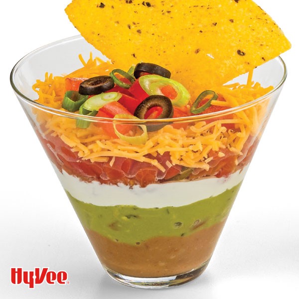 Glass cup layered with dips and topped with chopped tomatoes, shredded cheese, chopped green onions and tomatoes, sliced black olives with corn chip on top