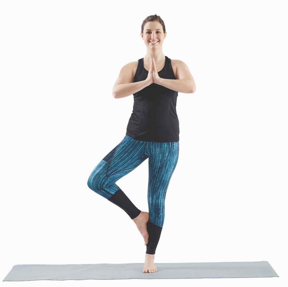 woman standing on her left foot with the bottom of the other foot pressed against the side of her left calf; her hands are in prayer position