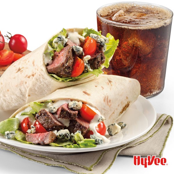Flour tortillas wrapped around lettuce, blue cheese, sliced steak, and halved cherry tomatoes with soda in a glass in backgroud